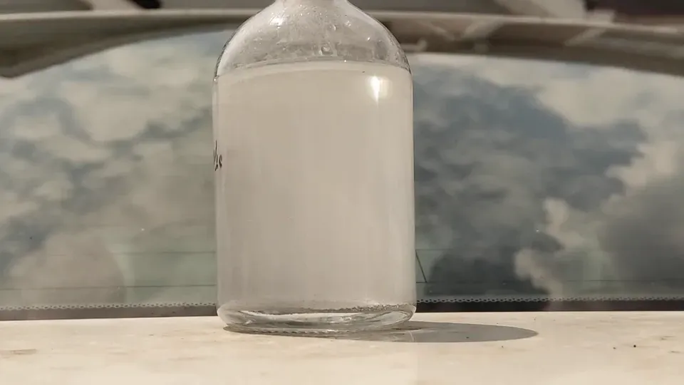 A stoppered glass bottle of cloudy liquid on a lab bench, backlit by a sunlit window.
